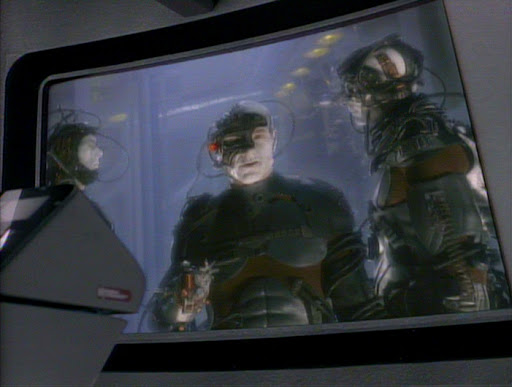 A screen-capture from Deep Space Nine pilot episode "The Emissary" showing Borg-assimilated Captain Picard (played by Patrick Stewart), covered in black mechanical prosthetics typical of the Borg. He is on the main view screen on the bridge of the USS Saratoga.